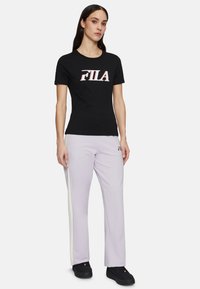 T-shirt noir à manches courtes avec logo "FILA" blanc et rouge, associé à un pantalon violet clair avec des bandes latérales. Des baskets noires complètent la tenue.