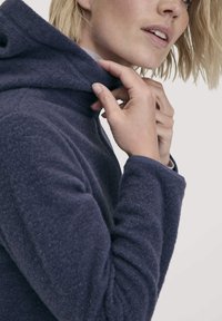 Marineblauwe fleece hoodie met een hoge kraag, gestructureerde stof en aansluitende mouwen. Een hand past de kraag aan, wat het zachte materiaal showt.