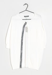 Biały dziergany kardigan z opuszczonymi ramionami, z kontrastową teksturowaną czarną lamówką wzdłuż przedniego otwarcia. Zawieszony na czarnym haczyku.