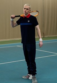 Tengerészkék polo vörös gallérral és kék csíkkal, sötét nadrággal párosítva. Teniszütőt tart a kezében, szürke és rózsaszín sportcipőt visel.