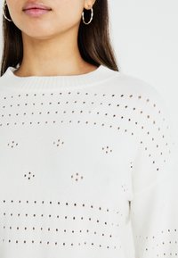 Pulover alb cu guler ribat, având un model texturat de mici găuri și puncte. Modelul poartă cercei rotunzi.