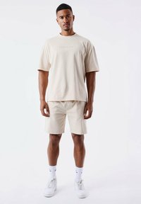 Camiseta de algodón beige con mangas cortas y cuello redondo, combinada con pantalones cortos beige a juego. Usada con zapatillas blancas y calcetines.