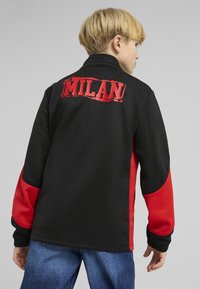 Sort og rød jakke med høj krave, struktureret sort stof og et iøjnefaldende rødt "AC MILAN!" logo prydet på ryggen.