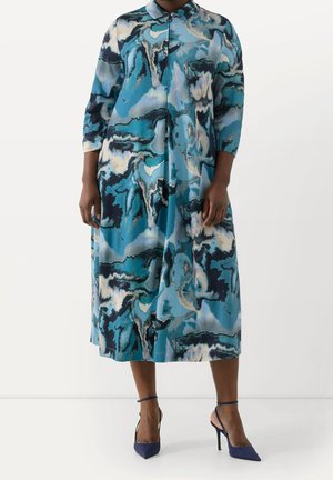 Femme portant une robe midi à manches longues, col bleu et noir à motif abstrait, avec des chaussures à talons hauts bleu marine à bride cheville.