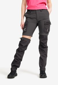 Person som bär svarta konvertibla cargo-byxor som delvis är dragkedjade på ett ben, i kombination med svarta stövlar och en rosa skjorta.