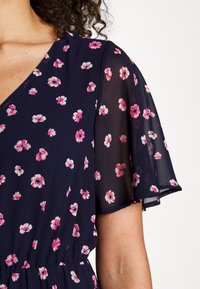 Femme portant une robe bleu marine à imprimé floral rose et blanc avec une manche légère et transparente.