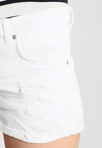 Pantalones cortos de mezclilla blanca con un diseño desgastado, con bordes deshilachados, dobladillo enrollado y cierre de botón metálico en la cintura.