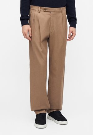 Pantaloni beige ampi indossati con una maglia nera a manica lunga e sneakers nere con suole bianche su sfondo bianco.