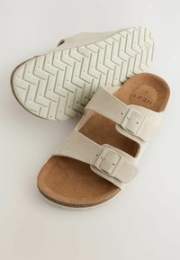 Beige ruskindsandaler med to justerbare stropper og en struktureret brun sål. Sålen har et fiskebensmønster for bedre greb.