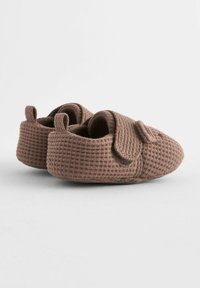 Papuci din bumbac maro, texturat, cu un model țesut, având un vârf rotunjit și o curea mică, înfășurată pe lateral pentru o purtare ușoară.