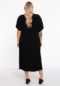 Robe midi noire à côtes avec des manches courtes, présentant une coupe décontractée et un dos ouvert. Portée avec des mules noires à talons carrés.
