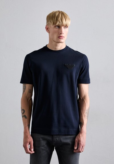 Navyblaue Kurzarm-T-Shirt aus weichem Baumwollstoff mit rundem Halsausschnitt und einem strukturierten schwarzen Logopatch auf der linken Brust.