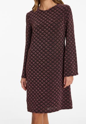 Kobieta nosząca sukienkę z długim rękawem sięgającą kolan z fakturowanym wzorem diamentu w czarnych i ciemnoczerwonych tonach.