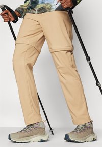 Beige színű, cipzáras lábszárú convertible nadrág, szürke túracipővel párosítva, amelyen lila részletek és megerősített talp található, trekking bottal tartva.