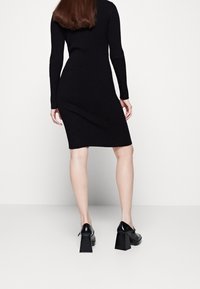 Černé žebrované pletené šaty s dlouhými rukávy, přiléhavý silueta, po kolena. Spárováno s lesklými černými botami na blokovém podpatku.