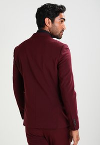 Burgundi színű, testre szabott zakó, amelynek strukturált kialakítása van, egysoros gombolással és fekete hajtókával. Sima textúrájú, négy gombos mandzsettával.