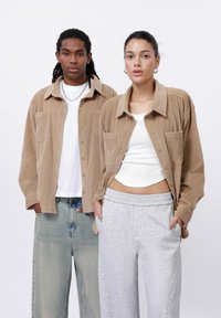 Dva modeli mají sladěné outfity. Mužský model má na sobě béžovou manšestrovou košili na knoflíky, světlé džíny a stříbrný řetízek. Ženský model nosí béžovou manšestrovou košili, bílý top na ramínka a šedé tepláky.