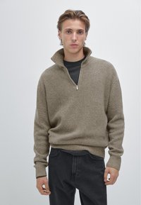 Béžový pletený svetr s vysokým límcem na zip, žebrovanou texturou a dlouhými rukávy, nošený přes tmavé tričko a doplněný tmavými džíny.