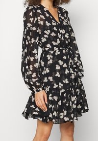 Černé květinové šaty s průhlednou tkaninou, dlouhými rukávy, výstřihem do V a vázáním v pase. Obsahují jemné květinové vzory a nařasený lem sukně.
