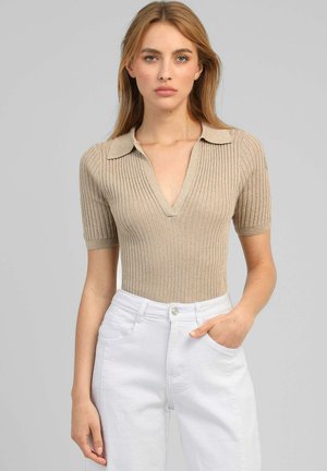 Jeune femme aux longs cheveux blonds porte un haut beige côtelé à manches courtes avec col et un pantalon blanc taille haute, main dans la poche.