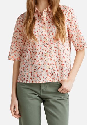 Kobieta nosząca krótką rękaw, zapinaną na guziki bluzkę z małym czerwono-różowym kwiatowym wzorem i oliwkowozielone spodnie, ręka w kieszeni.