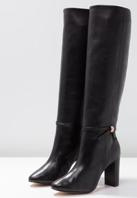 Knee-high black leather boots with a square toe and chunky heel. Features a decorative ribbon and a metallic accent near the ankle.