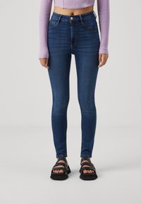 Kapeet tummansinisistä denim skinny-farkuista, joissa on korkea vyötärö, hienovaraiset ompeleet ja viiden taskun malli, yhdistettynä mustiin sandaaleihin.