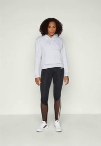 Sweatshirt cinza claro com capuz e superfície texturizada, combinado com leggings pretas com detalhes em malha e riscas laterais brancas, usado com ténis brancos.