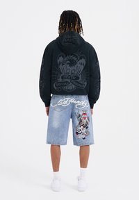 Felpa nera con grafica di un'aquila e testo sul retro; pantaloni corti in denim azzurro chiaro con ricami colorati di teschi e serpenti sui lati.