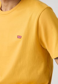 Man met een gele Levi's t-shirt met zichtbaar logo op de borst en een gedeeltelijke tatoeage op de arm, tegen een effen achtergrond.