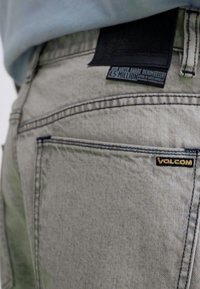 Light gray denim jeans featuring a black leather waistband label and a small yellow "VOLCOM" logo on the pocket, with visible stitching details.