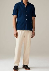 Chemise bleu marine à manches courtes avec un tissu texturé, associée à un pantalon beige à jambes larges et des mocassins foncés à semelle robuste.