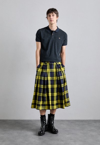 Dunkelblaues Polohemd mit Muster, kombiniert mit einem gelben und marineblauen Kilt mit Karomuster, ausgestattet mit Falten und seitlichen Taschen. Getragen mit glänzenden schwarzen Stiefeln.