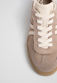 Baskets en daim beige avec des lacets blancs et des accents crème, vues de dessus sur un fond blanc.