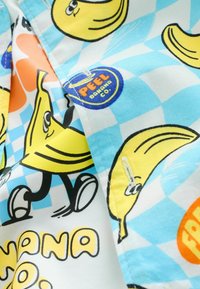 Buntes Stoffmuster mit gezeichneten Bananen, einige lächelnd und gehend, vor einem blau-weißen Karierten Hintergrund mit „PEEL“-Logo-Patches.