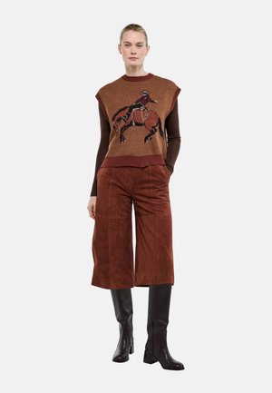 Femme portant un pull sans manches marron avec un motif cavalier à cheval, des culottes larges marron et des bottes noires montantes, debout devant un fond blanc.