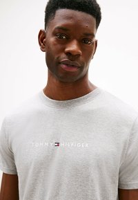 Szara bawełniana koszulka z okrągłym dekoltem, ozdobiona białym logo "TOMMY HILFIGER" oraz małym czerwonym i niebieskim akcentem.