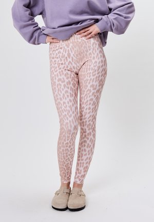 Person iført lyserøde og beige leopardmønstrede leggings, lilla sweatshirt og beige slip-on sko, står med hænderne på hoften.