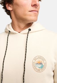 Sudadera color beige con cordón, que presenta un logo circular con un diseño de montaña y sol. Textura suave y corte casual, ideal para la comodidad.