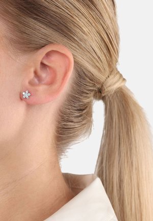 Acercamiento a la oreja de una mujer con un pequeño pendiente de diamante en forma de flor, pelo rubio recogido en una coleta y cuello de camisa blanca.