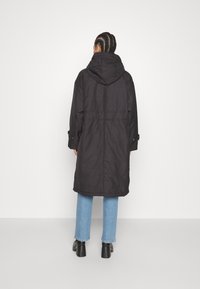 Manteau noir à capuche avec une coupe longue et décontractée, doté d'une taille cintrée et de fermetures à pression aux manches. Porté avec un jean bleu clair et des chaussures noires.