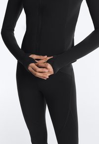 Person iført tætsiddende sort sportjakke og leggings, hænder foldet ved livet, tommelhuller synlige på jakkeærmerne.