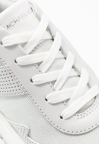 Bílé tenisky s texturovaným plátnovým svrškem, hladkými koženými akcenty a plochými bílými tkaničkami. Obsahují perforované detaily a jemné prošívání.