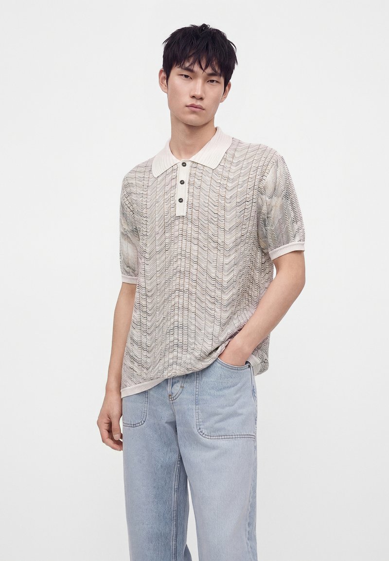 Joven con cabello corto y oscuro que lleva una camisa tipo polo de manga corta y estampada clara, y vaqueros azul claro con una mano en el bolsillo.
