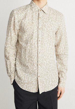 Mand iført en beige og lysebrun skjorte med bladmønster og knapper, samt sorte bukser mod en ensfarvet baggrund.