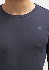 Mężczyzna ubrany w ciemnoniebieską, długą koszulkę z okrągłym dekoltem, z małym haftowanym logo "RAW" na lewej piersi.