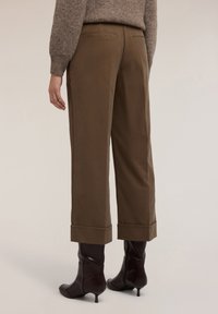 Person som bär högmidjade bruna vida byxor med uppvikta fållar och mörkbruna klackade ankelstövlar, mot en ljus bakgrund.