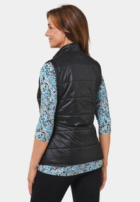Schwarze gesteppte Weste mit hohem Kragen, getragen über einem mehrfarbigen, gemusterten Langarmhemd. Die Seitenansicht zeigt das schlanke Design des Kleidungsstücks.