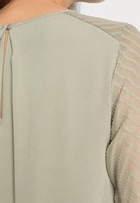 Blusa verde chiaro con chiusura a chiave sul retro, caratterizzata da maniche trasparenti a fantasia con design a zigzag e accenti testurizzati.