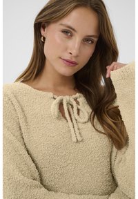 Beige textúrájú pulóver kerek nyakkivágással, masni részlettel és hosszú ujjal. Az anyag lágy és bolyhos hatású, kellemes érzetű.